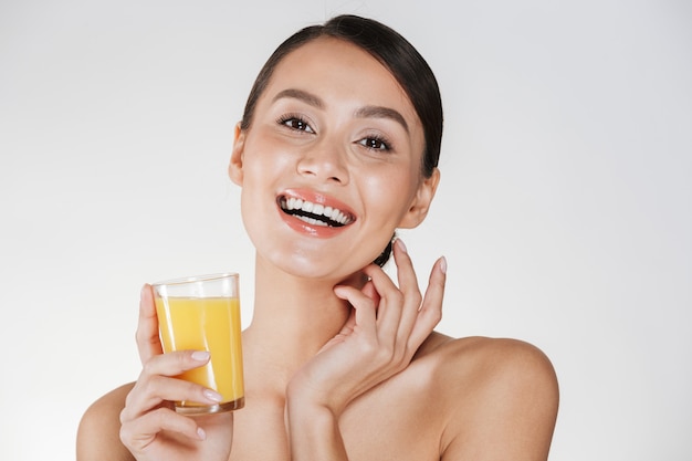 Счастливое изображение полуголой леди, улыбающейся и пьющей свежевыжатый апельсиновый сок из прозрачного стекла, изолированного над белой стеной