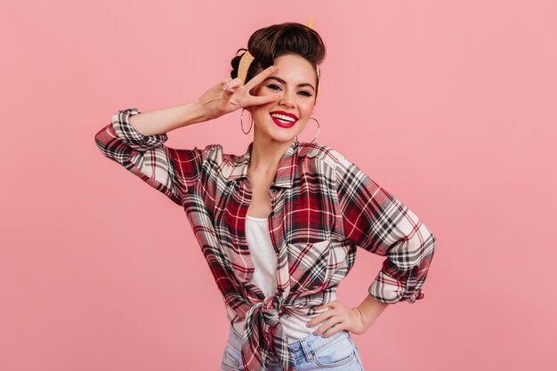 Happy pinup girl posing with peace sign. Studio shot of laughing beautiful woman in red checkered shirt.