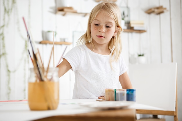 Happy, playful and cute blonde girl with freckles and beautiful blue eyes dressed in white t-shirt biting brush