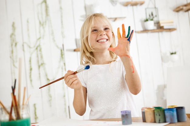 Happy and playful cute freckled blonde girl dressed in white, holding brush in one hand and showing another hand, which she messed up with paint.