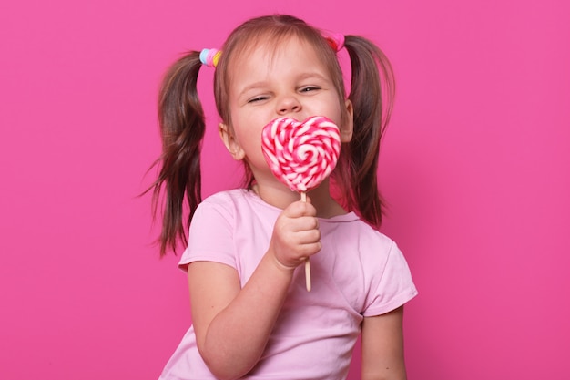 Happy playful cute girl wears rose t hirt, stands isolated on pink, holds bright lollipop in hand, eating it. Happy child enjoys spending free time. Childhood and emotions concept.