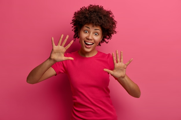 Happy playful young woman gestures with both hands, raises palms, shows ten gesture, laughs positively, wears rosy t shirt, stands indoor, has fun, eyes full of happines, foolishes around indoor