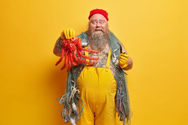 Счастливый, приятно удивленный бородатый боцман хвастается, что пойманного им большого осьминога держит в руке курительную трубку, в морском приключении, в желтом комбинезоне, с рыболовными снастями, в помещении, с большим толстым животом.