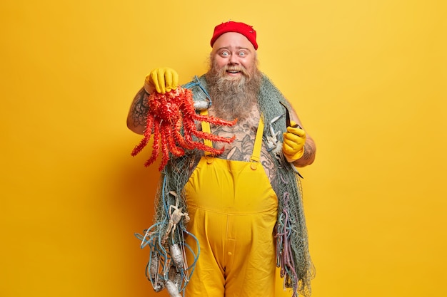 Happy pleasantly surprised bearded boatswain boasts what big octopus he caught holds smoking pipe has sea adventure wears yellow overalls carries fishing gears poses indoor has big fat belly