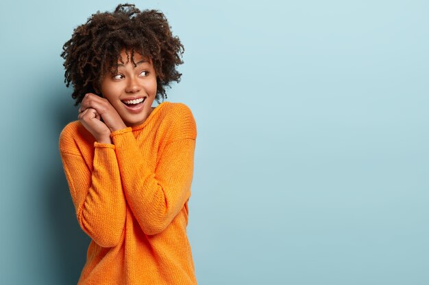 幸せな幸せなアフリカ系アメリカ人の女性は、顔の近くで手をつないで、焦点を合わせ、望ましいものに気づき、笑い、明るいセーターを着て、空きスペースを確保して青い壁に向かってポーズをとる