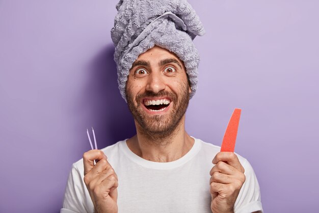 Happy pleased man with bristle holds nail file and tweezers, going to have beauty treatments in salon or at home, does nail up correction, has towel on head. Spa