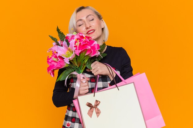 Счастливая и довольная молодая женщина в красивом платье, держащая букет цветов и бумажные пакеты с подарками, улыбается, празднует международный женский день, стоя над оранжевой стеной
