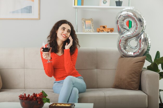 Happy and pleased young woman in casual clothes smiling cheerfully sitting on a couch with glass of wine talking on mobile phone in light living room celebrating international women's day march 8