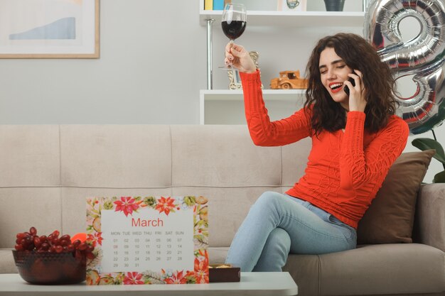 Счастливая и довольная молодая женщина в повседневной одежде, весело улыбаясь, сидит на диване с бокалом вина, разговаривает по мобильному телефону в светлой гостиной, празднуя международный женский день 8 марта