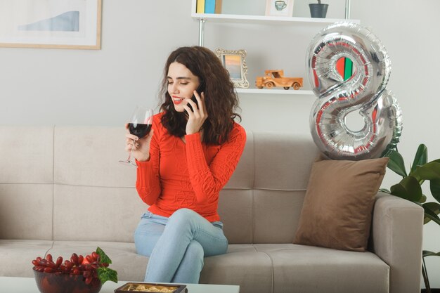 Happy and pleased young woman in casual clothes smiling cheerfully sitting on a couch with glass of wine talking on mobile phone in light living room celebrating international women's day march 8