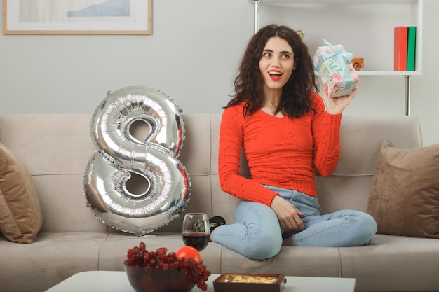 Happy and pleased young woman in casual clothes smiling cheerfully sitting on a couch with number eight shaped balloon holding present in light living room celebrating international womens day march 8