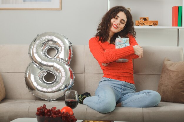 Happy and pleased young woman in casual clothes smiling cheerfully sitting on a couch with number eight shaped balloon hugging present celebrating international womens day march 8