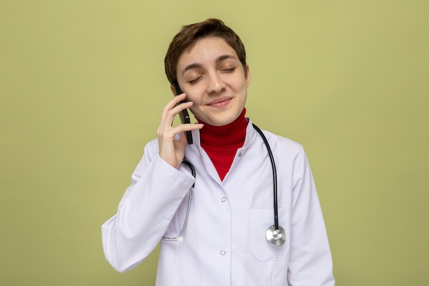 携帯電話で話している間自信を持って笑顔聴診器と白衣を着た幸せで喜んでいる若い女性医師
