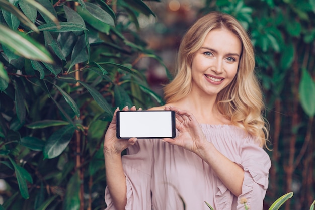 Happy portrait of a blonde young woman standing near the green plants showing mobile phone display