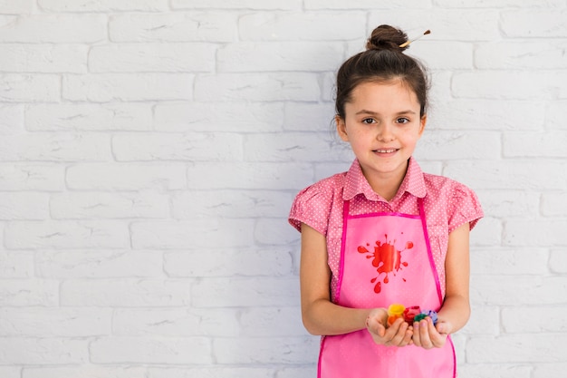 Happy portrait of a girl holding colorful paint bottles in hand standing against white wall