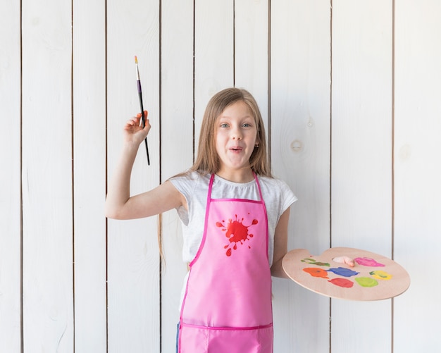 Free photo happy portrait of a girl holding paintbrush and wooden palette in hands standing against white wooden wall