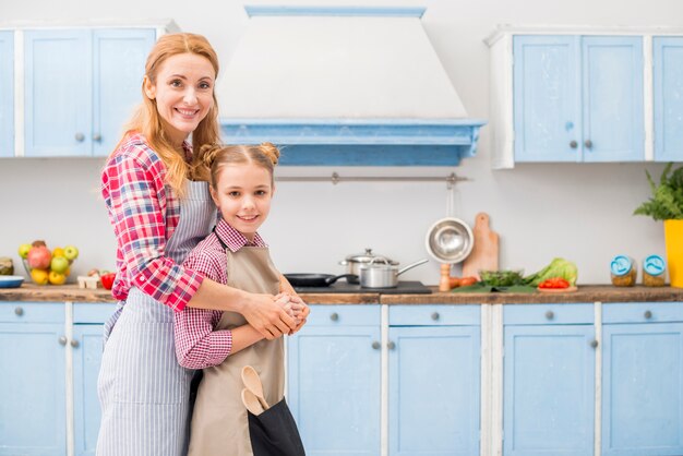 Счастливый портрет матери и дочери, глядя на камеру, стоя на кухне