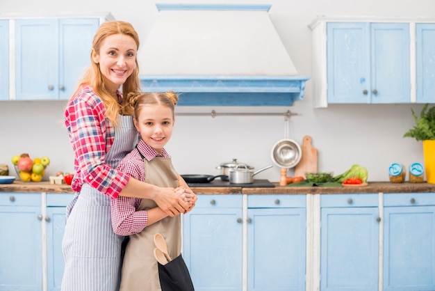 Happy portrait of mother and daughter looking at camera standing in the kitchen