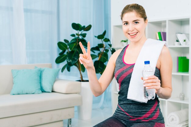 Счастливый портрет стройной молодой женщины, держащей бутылку с водой в руке, показываю знак победы