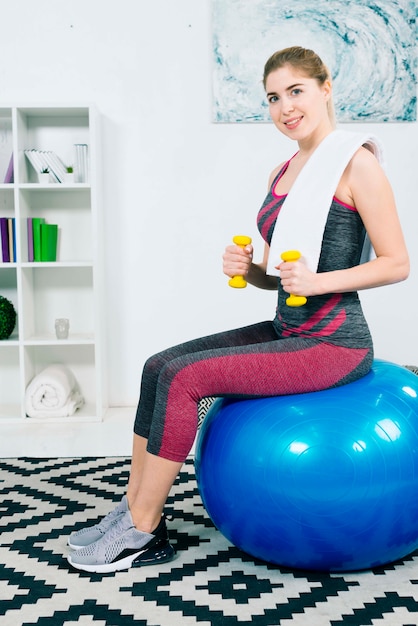 Счастливый портрет худой молодой женщины, сидящей на синем пилатесном мяче, тренирующемся с гантелями на ковре