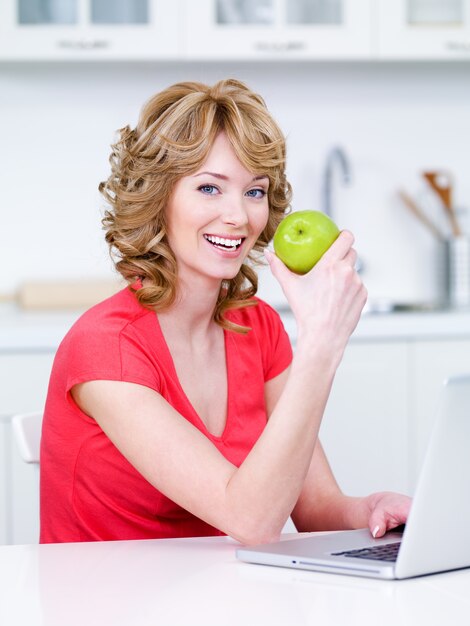 Happy portrait of young beautiful woman sitting in the kitchen and eating green apple