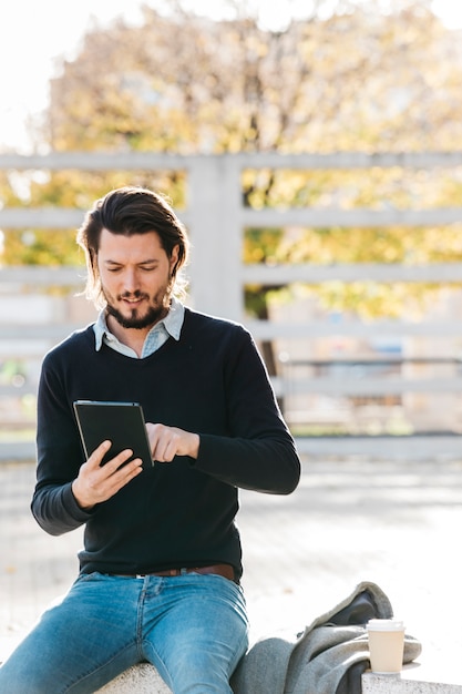 Happy portrait of a young man using smart phone sitting on bench with disposable coffee cup