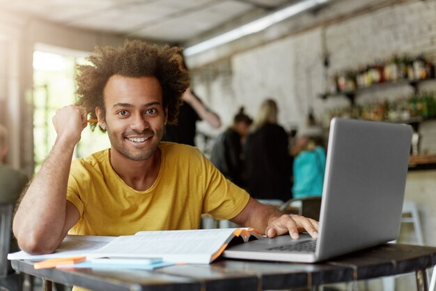 Happy positive African American college student with cheerful cute smile using wireless internet connection on laptop computer at coffee shop while looking for information online for research project