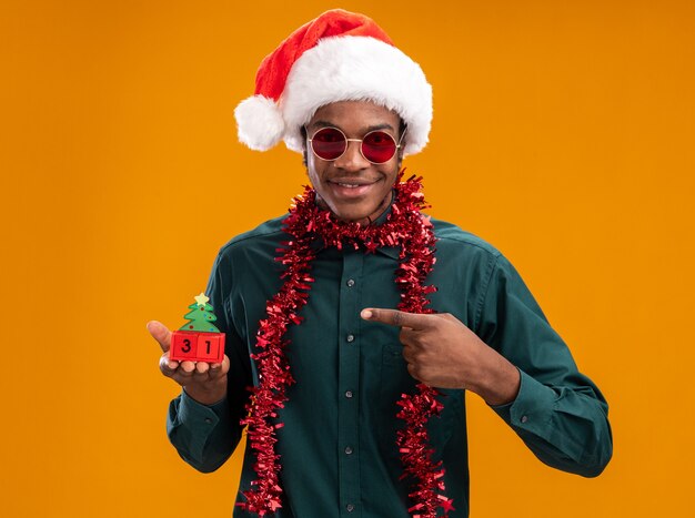 オレンジ色の背景の上に立って人差し指で指している新年の日付とおもちゃの立方体を保持しているサングラスを身に着けている花輪と花輪を持つサンタ帽子で幸せでポジティブなアフリカ系アメリカ人の男