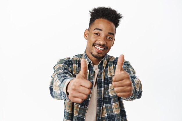 행복하고 긍정적인 아프리카계 미국인 남자가 엄지손가락을 치켜세우며 만족스럽게 웃으면서 훌륭한 일을 하고 있다고 말하며 배경 위에 서 있는 것을 격려합니다