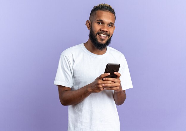 Happy and positive african american young man in white t-shirt holding smartphone looking at camera smiling cheerfully standing over blue background