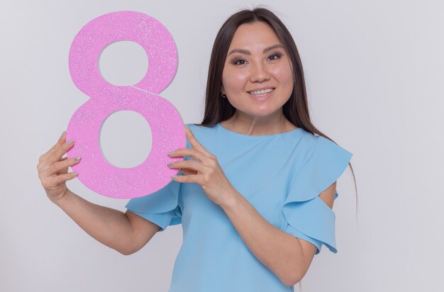 Счастливая и позитивная азиатская женщина, держащая номер восемь из картона, глядя вперед, весело улыбаясь, празднует международный женский день, стоя над белой стеной