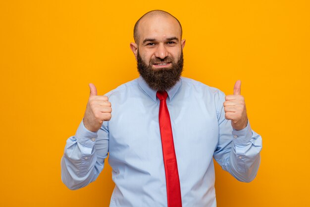 Happy and positive bearded man in red tie and shirt looking at camera smiling cherfully showing thumbs up standing over orange background