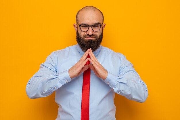 Happy and positive bearded man in red tie and shirt wearing glasses holding hands together waiting for something standing over orange background