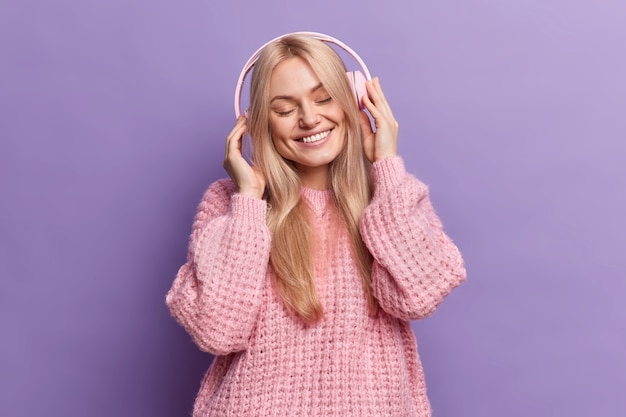 Happy positive blonde womann closes eyes and smiles with satisfaction listens audio track via headphones dressed in casual knitted jumper 
