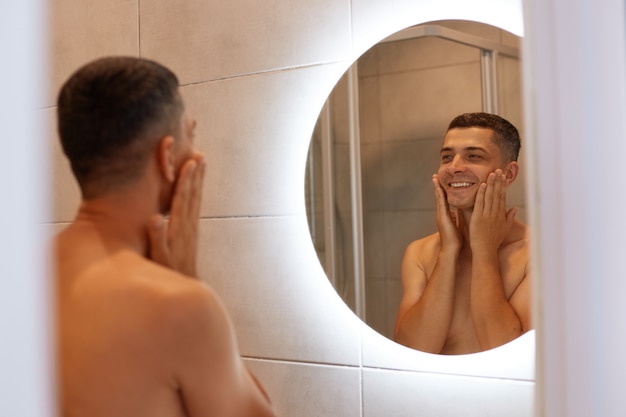 Happy positive brunette man standing in bathroom, looking at his reflection in the mirror, touching his cheeks, applying shaving agent on face, smiling.