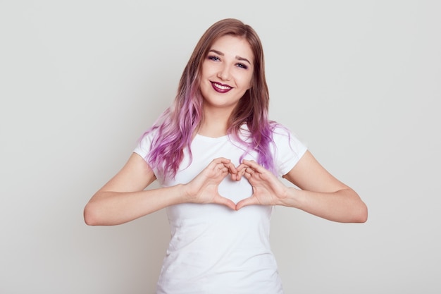 Happy positive female with bright lilac hair with charming smile and showing heart gesture with fingers, expressing love, posing isolated over gray wall.