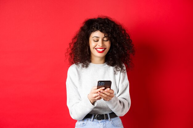 Happy positive girl chatting on phone, reading message and smiling, using social media app, standing on red background.