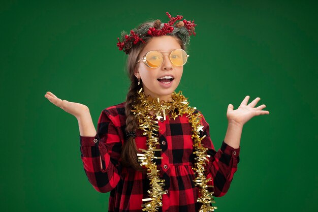 목 주위에 반짝이와 체크 드레스에 크리스마스 화환을 입고 행복하고 긍정적 인 어린 소녀 행복하고 쾌활한