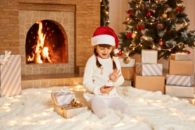 Happy positive little girl wearing white sweater and santa claus hat, sitting on floor near Christmas tree, present boxes and fireplace, having video call with friends via smartphone.