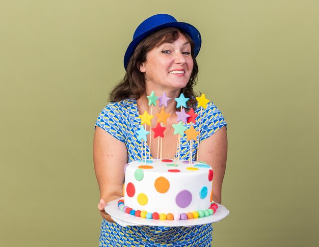 Happy and positive middle age woman in party hat holding birthday cake  smiling cheerfully celebrating birthday party standing over green wall