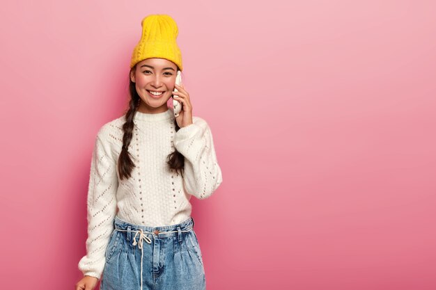 Happy positive mixed race teenage woman enjoys communication via cellphone, wears stylish yellow hat