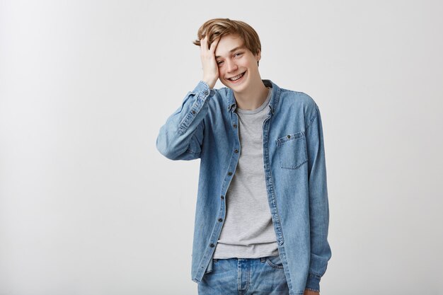 Happy positive pleasant looking male model in denim shirt and jeans, with fair hair and blue eyes, smiles broadly, feels a little bit shy, touches his hair. Beauty and youth concept