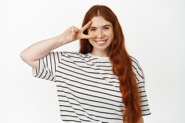 Happy and positive redhead girl showing peace sign near clean no makeup skin, v-sign on eye, standing cheerful in t-shirt on white studio.