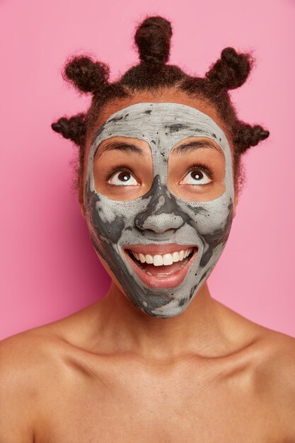 Happy positive woman looks upwards, applies purifying facial mask, removes blackheads, looks curiously upwards, stands shirtless, has well cared body healthy skin combed hair buns poses over rosy wall