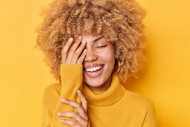 Happy positive woman with curly bushy hair makes face palm smiles broadly shows white teeth keeps eyes closed wears comfortable sweater isolated over vivid yellow background Sincere feelings