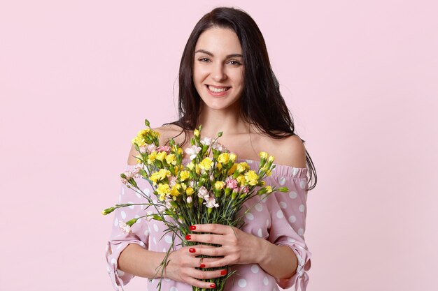 Happy positive woman with dark hair, holds flowers in hands, smiles positively, enjoys spring warm day, dressed in stylish polka dot dress