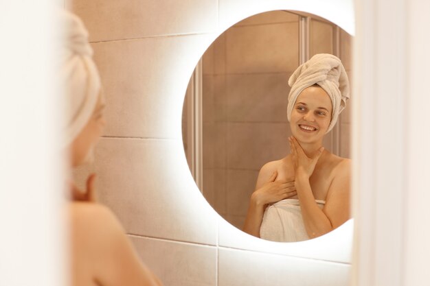 Happy positive woman with fresh skin looking on reflection in the mirror after shower, standing wrapped in white towel, posing in bathroom while doing morning procedures.