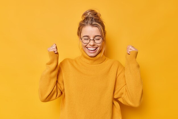 お団子に集まった髪の幸せなポジティブな女性拳は成功を喜ぶ勝利を祝うか、素晴らしい結果は黄色の壁に対してカジュアルなジャンパーラウンド眼鏡ポーズを着ますはい私はついにそれをしました