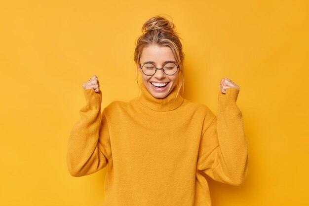 Happy positive woman with hair gathered in bun clenches fists rejoices success celebrates victory or great result wears casual jumper round spectacles poses against yellow wall Yes I finally did it