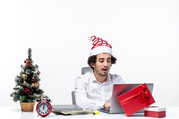 Happy positive young businessman with funny santa claus hat looking at something on laptop in the office on white background